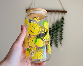 May include: A clear glass reusable water bottle with a bamboo lid and straw. The bottle has a lemon print design with yellow lemons and green leaves.