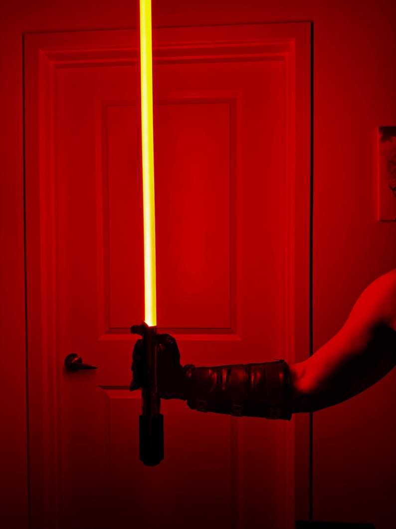 May include: A black lightsaber with a bright yellow blade is held in front of a red door. The hilt is black, and the blade extends vertically. The scene is illuminated with red light, creating a dramatic effect.
