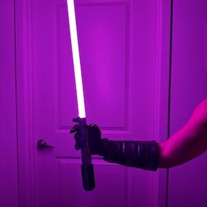 May include: A purple lightsaber with a bright white blade is held in a black gloved hand. The lightsaber has a black handle. The background is a purple-lit room with a white door.