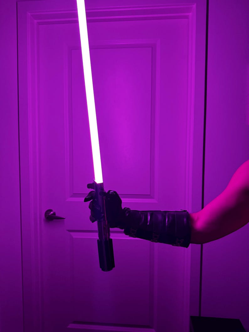 May include: A purple lightsaber with a bright white blade is held in a black gloved hand. The lightsaber has a black handle. The background is a purple-lit room with a white door.