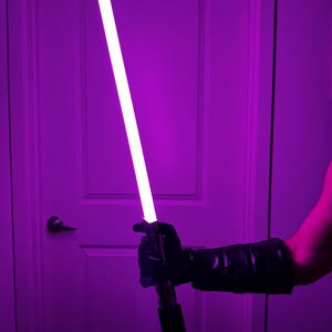 May include: A purple lightsaber with a bright white blade is held in a black gloved hand. The lightsaber has a black and silver handle. The background is a purple-lit room with a white door.