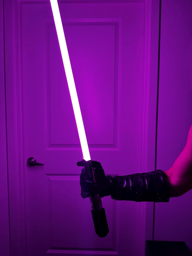May include: A purple lightsaber with a bright white blade is held in a black gloved hand. The lightsaber has a black and silver handle. The background is a purple-lit room with a white door.