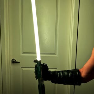 May include: A silver and black lightsaber with a bright white blade is held by a person wearing a black glove. The lightsaber is illuminated, and the background is a white door.
