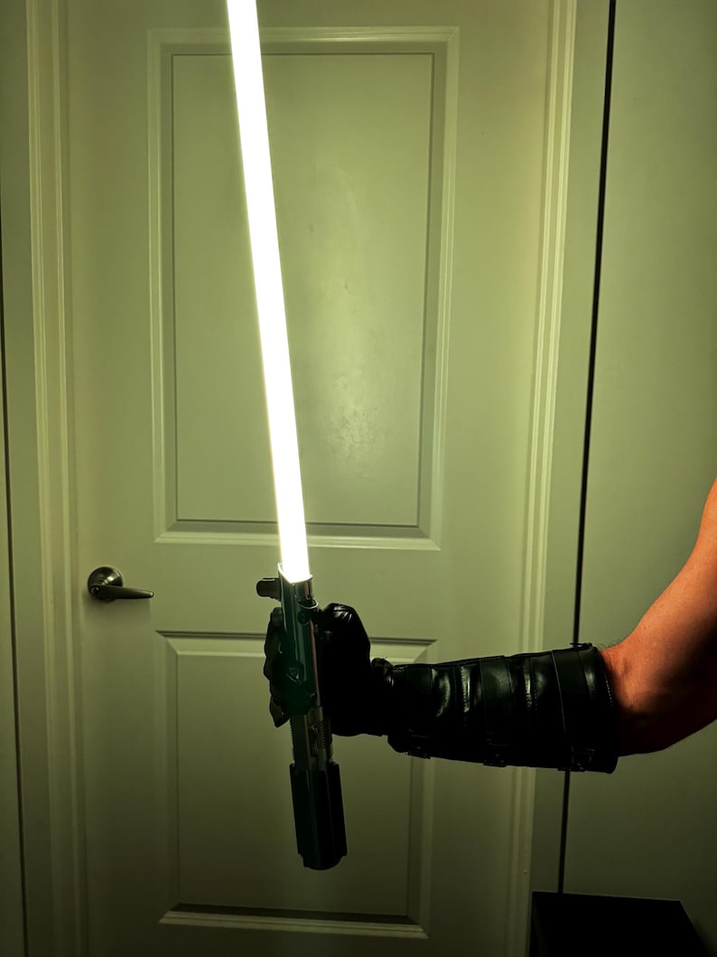 May include: A silver and black lightsaber with a bright white blade is held by a person wearing a black glove. The lightsaber is illuminated, and the background is a white door.