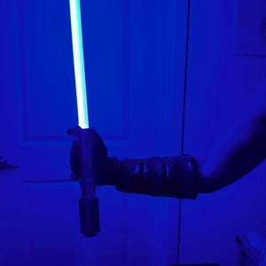 May include: A blue lightsaber with a glowing blade is held in a gloved hand. The hilt is dark, and the blade emits a bright, cool light. The background is a deep blue.