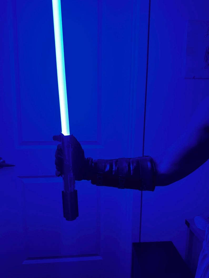 May include: A blue lightsaber with a glowing blade is held in a gloved hand. The hilt is dark, and the blade emits a bright, cool light. The background is a deep blue.