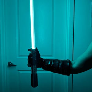May include: A black lightsaber with a bright blue blade is held in a gloved hand. The lightsaber is against a teal background, with a white door and a doorknob visible.