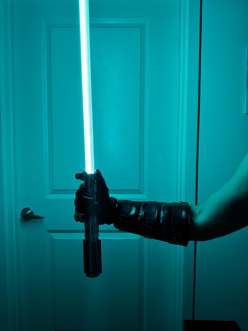 May include: A black lightsaber with a bright blue blade is held in a gloved hand. The lightsaber is against a teal background, with a white door and a doorknob visible.
