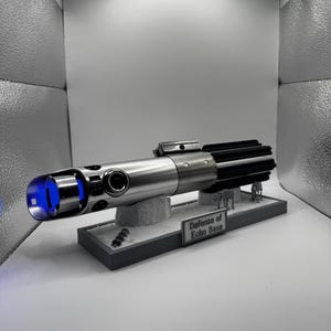 Könnte beinhalten: Silberfarbene Lichtschwert-Replik mit schwarzem Griff und blau leuchtender Klinge. Das Lichtschwert ruht auf einer Ausstellungsbasis mit dem Text "Defense of Echo Base". Die Basis hat kleine Figuren.