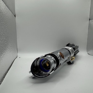 May include: A silver lightsaber hilt with a black emitter and gold accents. The hilt has a cylindrical design with a blue inner component and a brown button. The lightsaber is a prop from a science fiction franchise.