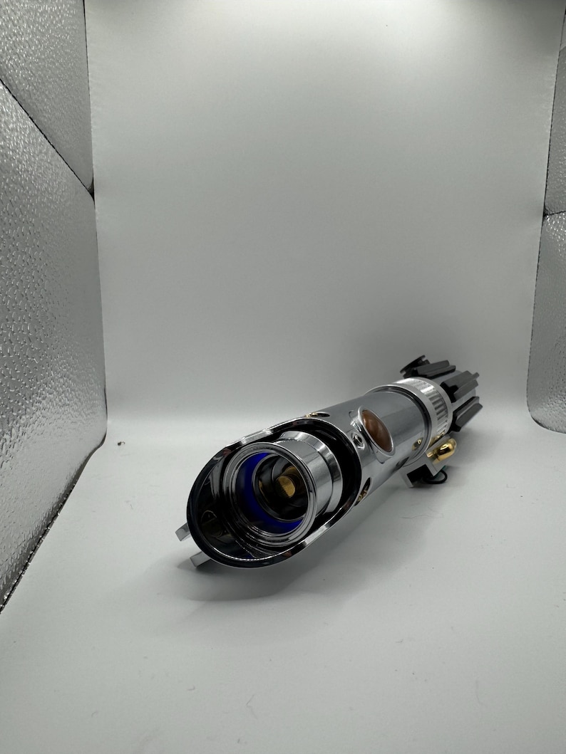 May include: A silver lightsaber hilt with a black emitter and gold accents. The hilt has a cylindrical design with a blue inner component and a brown button. The lightsaber is a prop from a science fiction franchise.