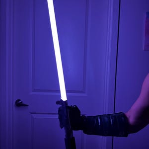 May include: A glowing white and purple lightsaber is held in a black gloved hand. The lightsaber has a black handle and a bright white blade with a purple hue. The background is a dimly lit room with a white door.