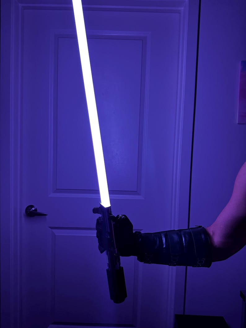 May include: A glowing white and purple lightsaber is held in a black gloved hand. The lightsaber has a black handle and a bright white blade with a purple hue. The background is a dimly lit room with a white door.