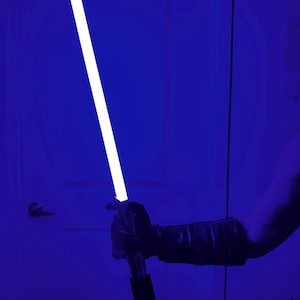 May include: A blue lightsaber with a bright white blade is held against a blue background. The hilt is black and silver. The person holding the lightsaber is wearing a black glove and sleeve.