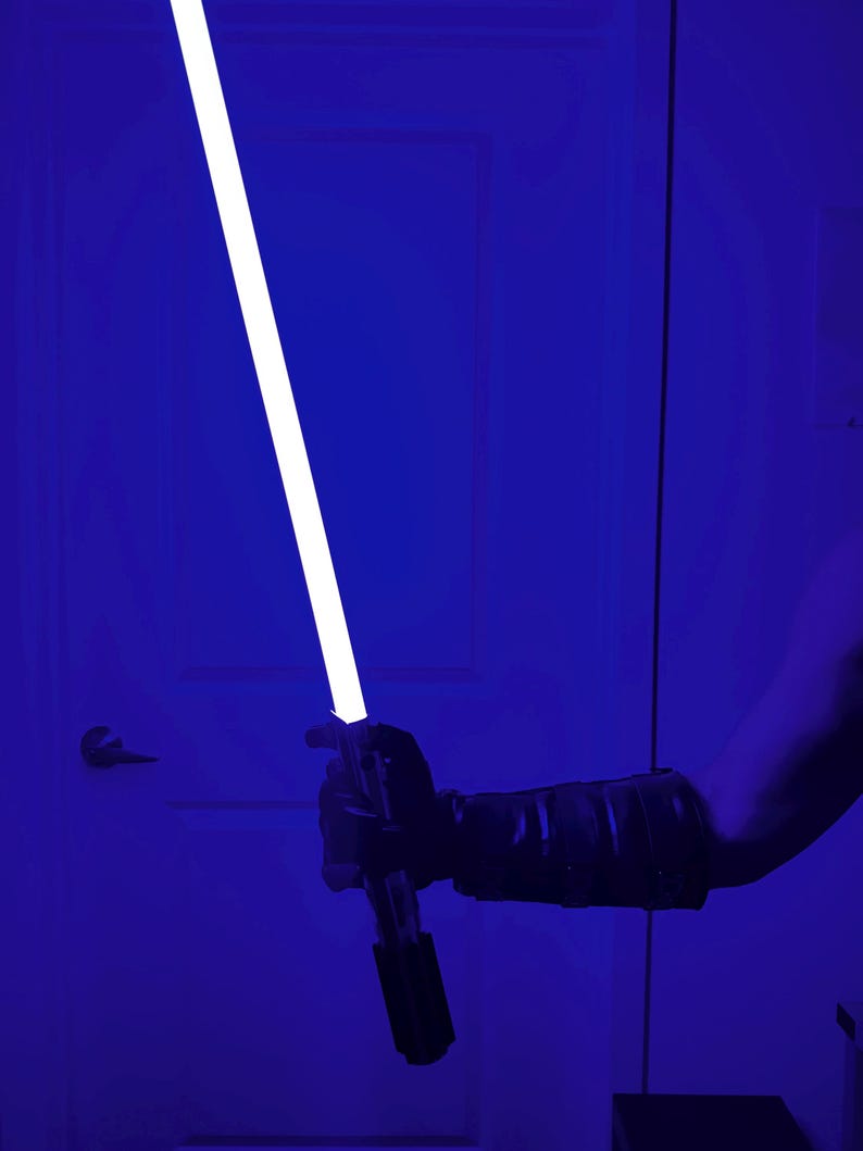 May include: A blue lightsaber with a bright white blade is held against a blue background. The hilt is black and silver. The person holding the lightsaber is wearing a black glove and sleeve.