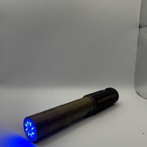 May include: A cylindrical flashlight with a bronze-colored body and a black head. The flashlight emits a bright blue light from the front, illuminating the surrounding area. The light source is composed of multiple small LEDs.