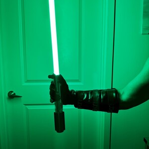 May include: A green lightsaber with a bright, glowing blade. The hilt is black and silver, held by a person wearing a black glove and arm guard. The background is a green-tinted room.