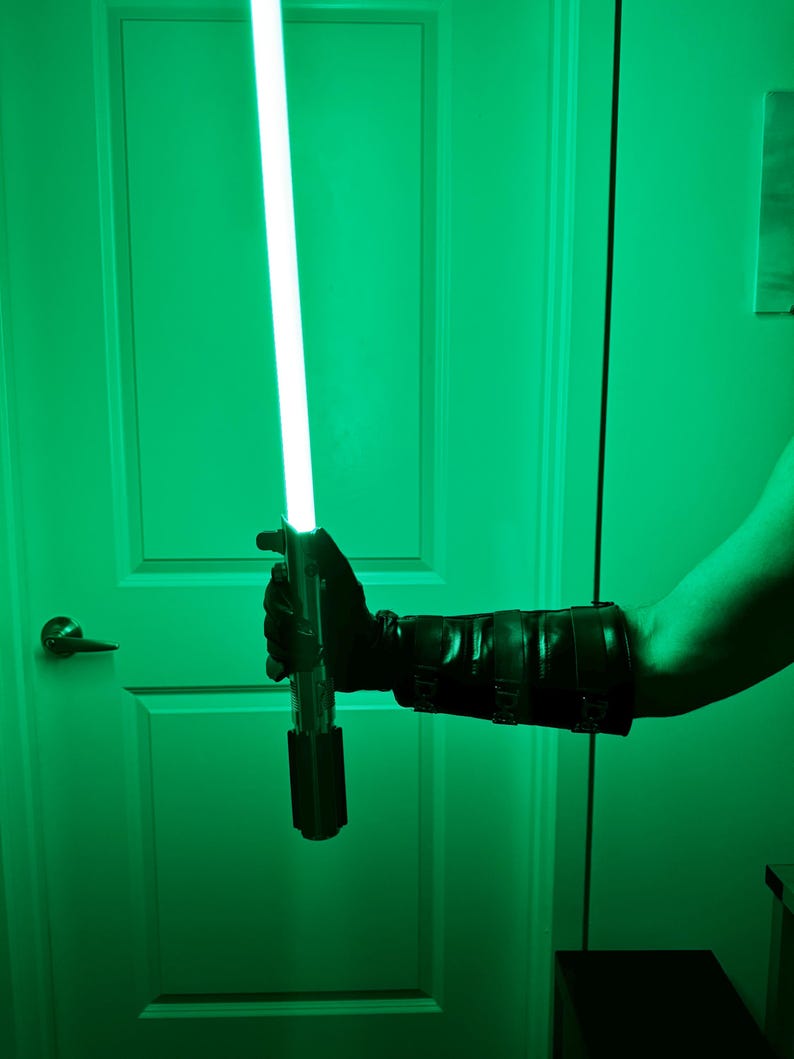 May include: A green lightsaber with a bright, glowing blade. The hilt is black and silver, held by a person wearing a black glove and arm guard. The background is a green-tinted room.