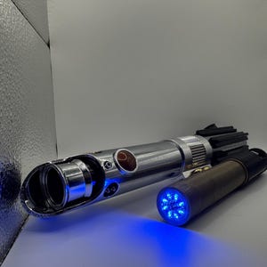 May include: Two metallic lightsabers with blue LED lights. One is silver with a black handle, the other is brown and black. The lightsabers are on a white surface.