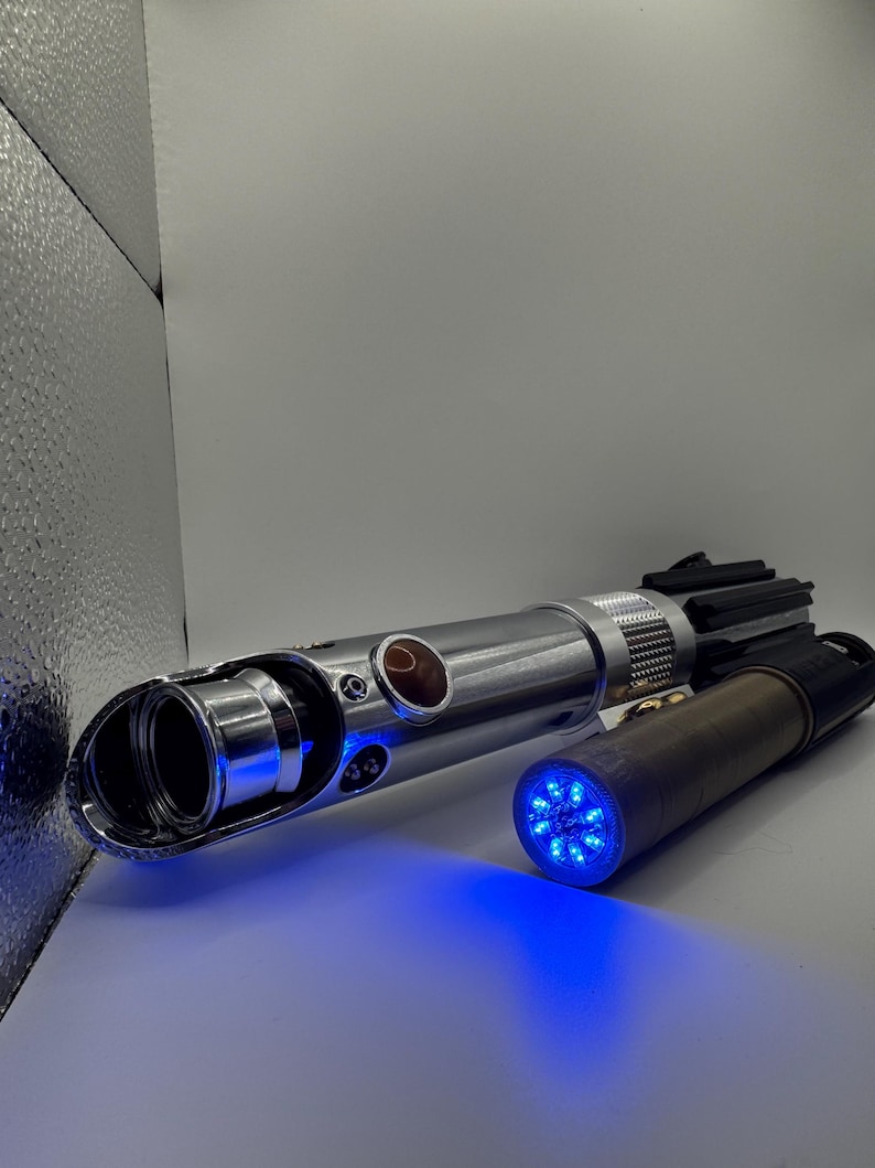 May include: Two metallic lightsabers with blue LED lights. One is silver with a black handle, the other is brown and black. The lightsabers are on a white surface.