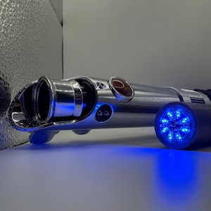May include: A silver lightsaber with a blue glowing end and a blue light emanating from the handle. The lightsaber has a circular red button and two black buttons. The lightsaber is on a white surface.