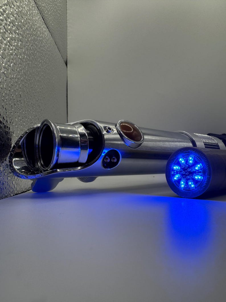 May include: A silver lightsaber with a blue glowing end and a blue light emanating from the handle. The lightsaber has a circular red button and two black buttons. The lightsaber is on a white surface.