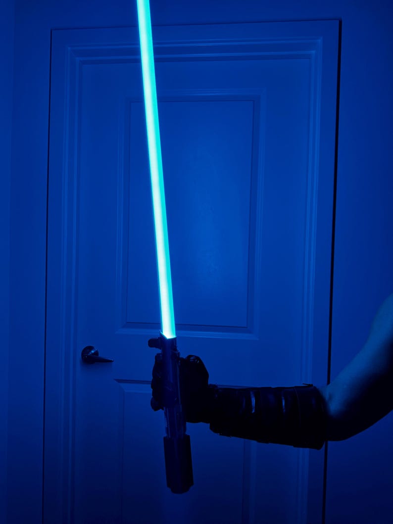 May include: A blue lightsaber with a bright, glowing blue blade is held in a gloved hand. The lightsaber is positioned in front of a white door, creating a dramatic, sci-fi themed image.