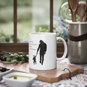 Peut inclure: Mug en céramique blanche avec une silhouette noire et blanche d'un parent et d'un enfant se tenant la main. Le mug porte le texte "Mit Papa an meiner Seite gibt es keine Grenzen". Le mug est posé sur une planche à découper en bois.