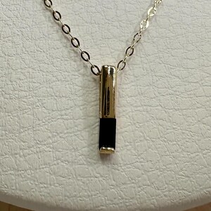 May include: A gold chain necklace with a black and gold pendant. The pendant is a long, thin rectangle with a black stone in the center.
