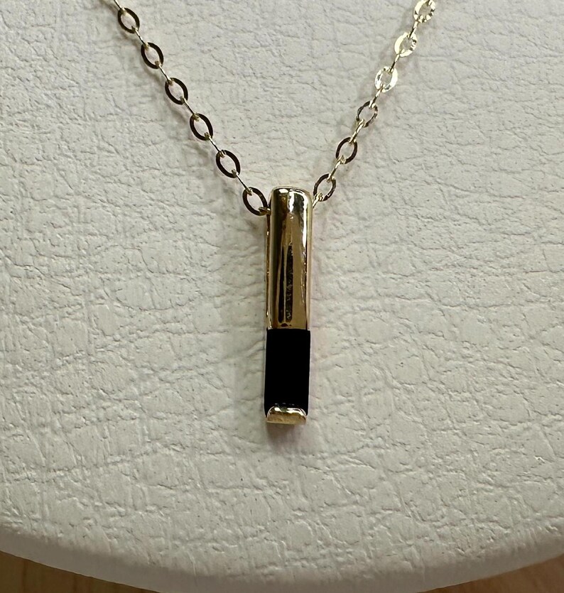 May include: A gold chain necklace with a black and gold pendant. The pendant is a long, thin rectangle with a black stone in the center.
