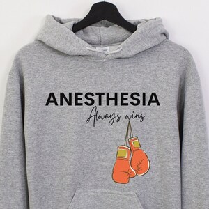 May include: A gray hooded sweatshirt with the text "Anesthesia Always Wins" and a graphic of two red boxing gloves hanging from a string.