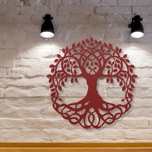 May include: A red wooden tree of life wall decor with intricate details and a circular shape. The tree is mounted on a white brick wall with two black wall lights on either side.