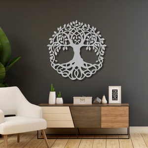 May include: A silver metal tree of life wall decor with intricate branches and roots. The decor is hanging on a dark grey wall in a living room setting with a white armchair and a wooden console table.