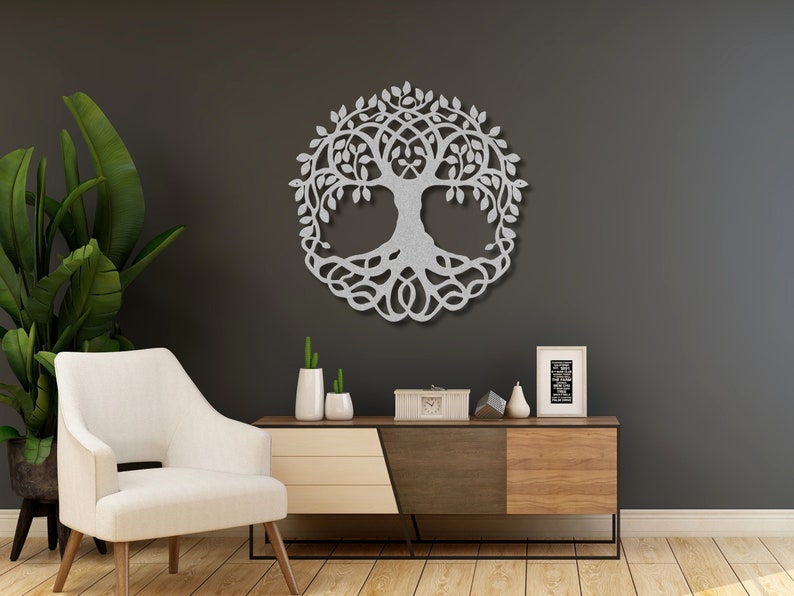 May include: A silver metal tree of life wall decor with intricate branches and roots. The decor is hanging on a dark grey wall in a living room setting with a white armchair and a wooden console table.