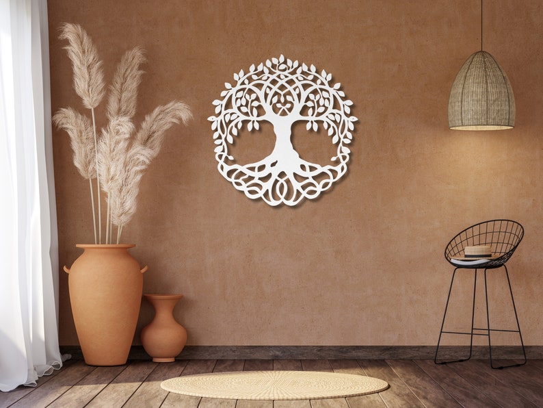 May include: White wooden wall decor in the shape of a tree of life with intricate branches and roots. The decor is hanging on a brown wall with a vase of dried pampas grass and a black metal stool in the foreground.