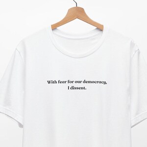 May include: White t-shirt with the text "With fear for our democracy, I dissent."