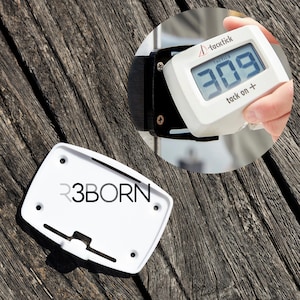 Puede incluir: Soporte de plástico blanco con el texto "R3BORN" impreso en él. Un temporizador digital blanco con una correa negra está unido al soporte. El temporizador muestra "309" y el texto "tack on +" en la pantalla.