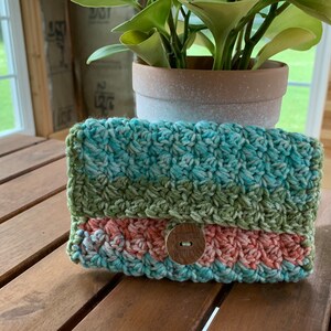 May include: A handmade crochet wallet in shades of teal, green, and coral. The wallet features a textured stitch pattern and a wooden button closure with the words "handmade with love" engraved on it. The wallet is sitting on a wooden surface.
