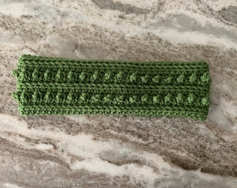 May include: A green crocheted headband with a textured pattern. The headband is made with a thick yarn and has a simple design.