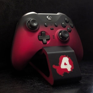 May include: A red and black Xbox controller resting on a black stand. The controller has a gradient design, transitioning from black to red. The stand features a white number 4 on a red splatter design.