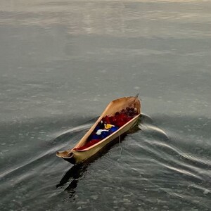 K&ouml;nnte beinhalten: Ein kleines, h&ouml;lzernes Boot schwimmt auf dem Wasser. Das Boot ist gef&uuml;llt mit roten, blauen und gelben Gegenst&auml;nden. Das Wasser ist ruhig und der Himmel ist hellblau.