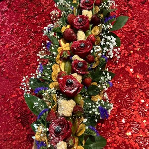 K&ouml;nnte beinhalten: Ein langes Holztablett gef&uuml;llt mit einer Vielzahl von Fr&uuml;chten, Blumen und anderen Dekorationselementen. Das Tablett ist mit roten Rosen, Erdbeeren, Trauben und wei&szlig;en Blumen verziert. Das Tablett befindet sich auf einer roten, paillettenbesetzten Tischdecke.