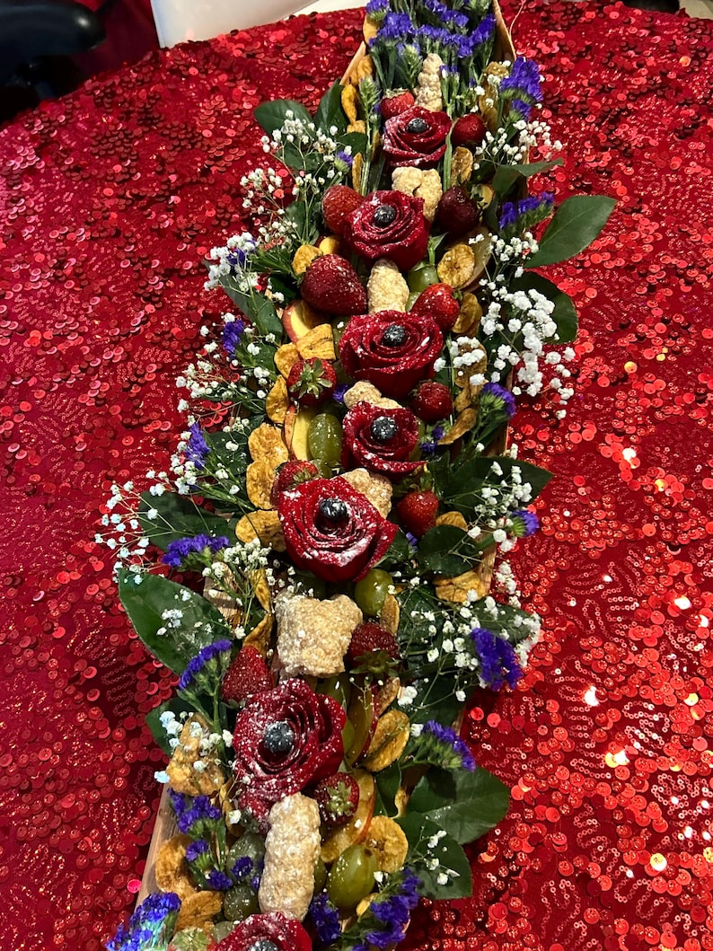 K&ouml;nnte beinhalten: Ein langes Holztablett gef&uuml;llt mit einer Vielzahl von Fr&uuml;chten, Blumen und anderen Dekorationselementen. Das Tablett ist mit roten Rosen, Erdbeeren, Trauben und wei&szlig;en Blumen verziert. Das Tablett befindet sich auf einer roten, paillettenbesetzten Tischdecke.