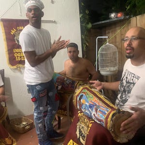 Puede incluir: Tres personas están reunidas alrededor de un tambor grande y decorado. El tambor está cubierto con una tela marrón con ribetes dorados y un diseño colorido. La persona de la izquierda lleva una camisa blanca, pantalones vaqueros azules y un gorro de cocinero blanco. La persona del medio está sin camisa y tiene el pelo corto castaño. La persona de la derecha lleva una camiseta blanca con un gráfico negro y gafas con montura negra. El texto en la pancarta detrás de las personas dice "AÑÁ OBB ENRIQ OGE 15-10".