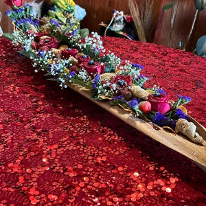 K&ouml;nnte beinhalten: Ein langes Holztablett gef&uuml;llt mit roten Rosen, Blaubeeren und anderen getrockneten Fr&uuml;chten und Blumen. Das Tablett steht auf einer roten, paillettenbesetzten Tischdecke.