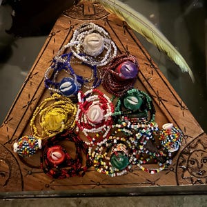 Puede incluir: Una bandeja triangular de madera con un diseño de sol tallado, que contiene nueve collares de cuentas de colores con conchas de cauri. Cada collar tiene un esquema de color y un patrón diferente.