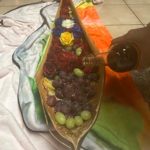 K&ouml;nnte beinhalten: Eine h&ouml;lzerne, bootf&ouml;rmige Servierplatte gef&uuml;llt mit roten, gelben und blauen Rosen, gr&uuml;nen Trauben und wei&szlig;en Streuseln.