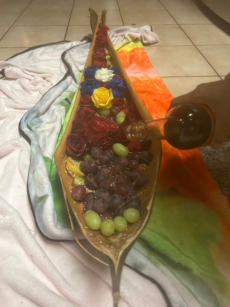 K&ouml;nnte beinhalten: Eine h&ouml;lzerne, bootf&ouml;rmige Servierplatte gef&uuml;llt mit roten, gelben und blauen Rosen, gr&uuml;nen Trauben und wei&szlig;en Streuseln.