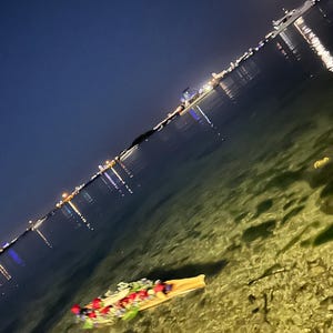K&ouml;nnte beinhalten: Ein mit roten, wei&szlig;en und blauen Blumen verziertes Holzboot schwimmt nachts in klarem, flachem Wasser. Das Boot wird vom Mond und den in das Wasser reflektierten Lichtern der Stadt beleuchtet.