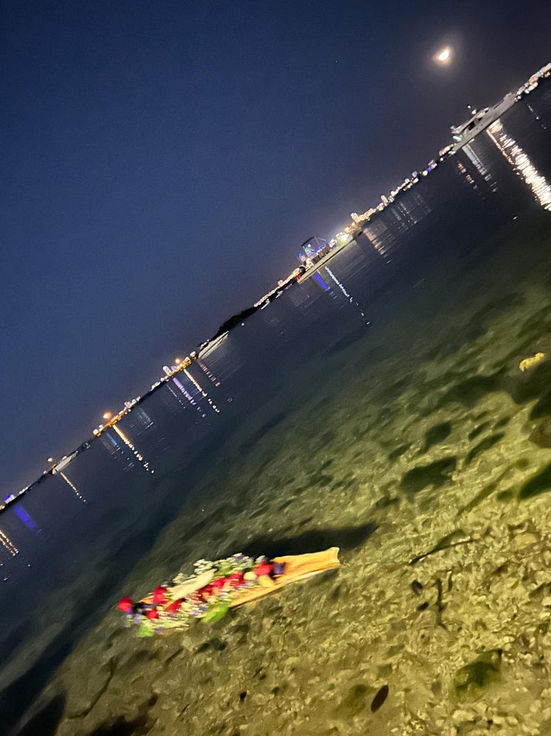 K&ouml;nnte beinhalten: Ein mit roten, wei&szlig;en und blauen Blumen verziertes Holzboot schwimmt nachts in klarem, flachem Wasser. Das Boot wird vom Mond und den in das Wasser reflektierten Lichtern der Stadt beleuchtet.